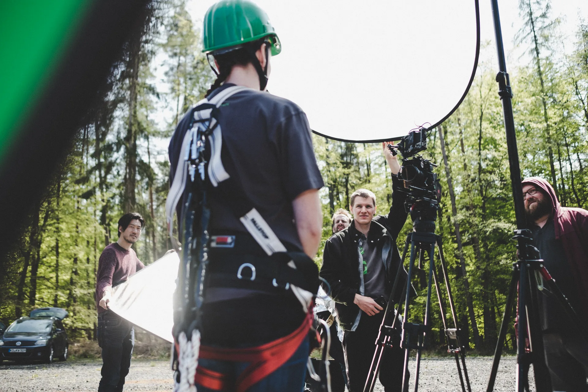 Filmteam bei Außenaufnahmen im Wald, mit Kamera auf Stativ, Reflektor und Personen in Arbeitskleidung.