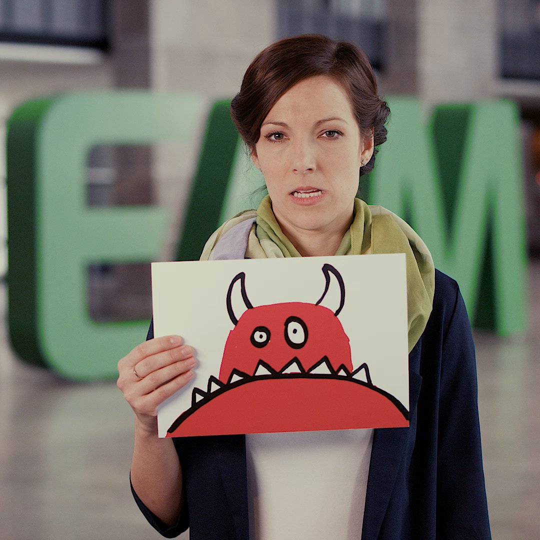 Eine Frau hält ein Bild mit einem roten Monster, im Hintergrund sind große grüne Buchstaben EAM zu sehen.