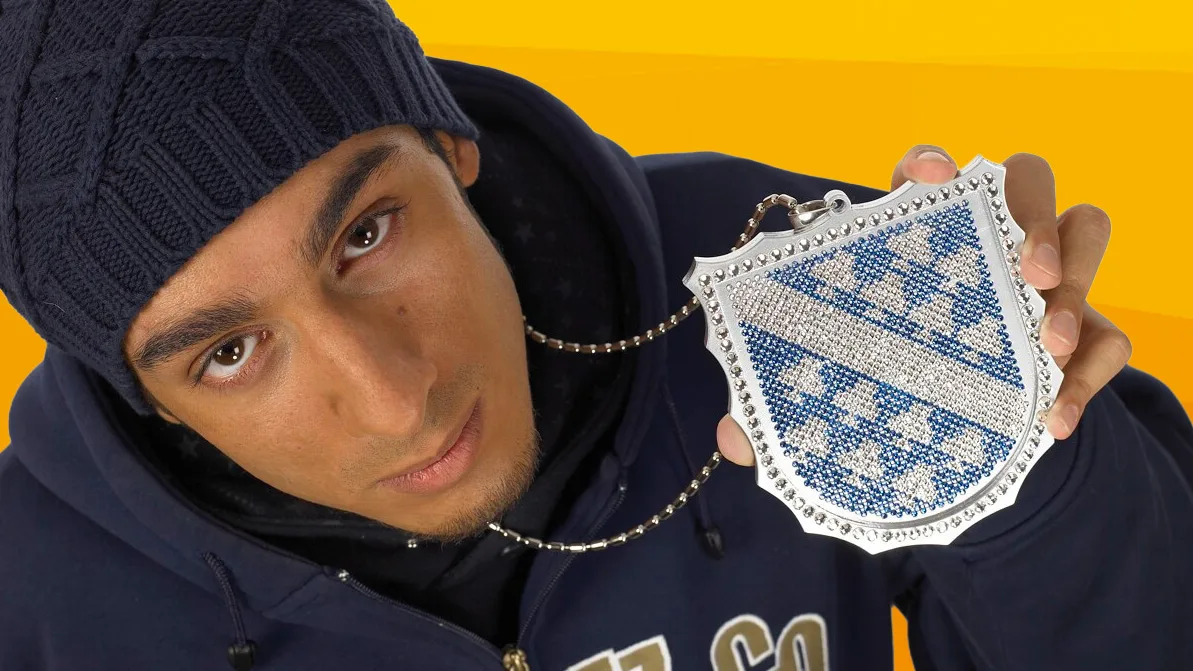 Ein junger Mann mit dunkelblauer Strickmütze und Kapuzenpullover hält eine glitzernde, Kette mit dem Kassel-Wappen in die Kamera.